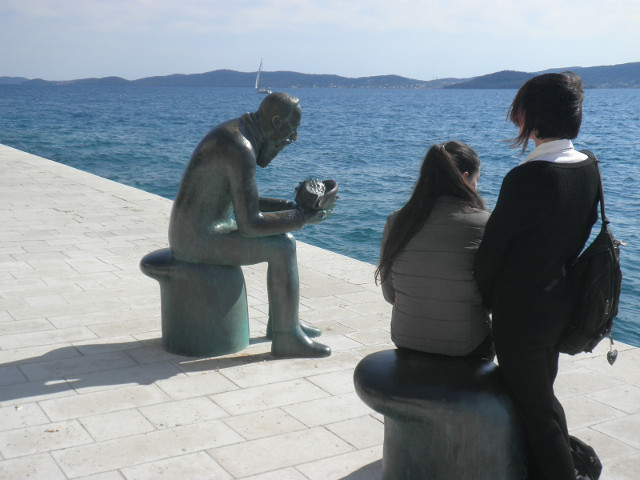 living and frozen tourists observing a metal statue of sitting man studying an object in his hands